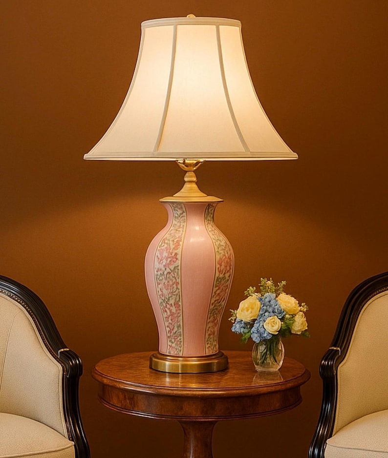 May include: A pink ceramic table lamp with a floral design, a cream-colored lampshade, and a brass base. The lamp sits on a round wooden table, next to a small vase of flowers. Two chairs are on either side of the table.