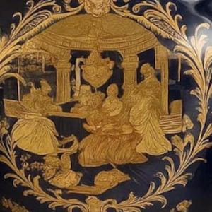 Può includere: Un vaso di ceramica blu scuro con dettagli in rilievo dorati ornati. Il rilievo raffigura una scena classica con figure sotto una struttura ad arco, circondata da motivi floreali e volute. Il vaso ha una finitura lucida.