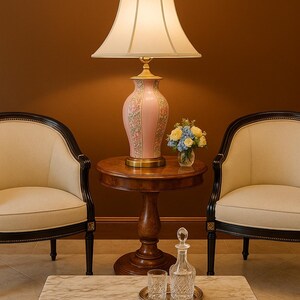 May include: A pink floral table lamp with a cream-colored lampshade sits on a small wooden table. Two cream-colored chairs with black trim flank the table. A marble-topped coffee table holds a decanter, two glasses, and a small tray.