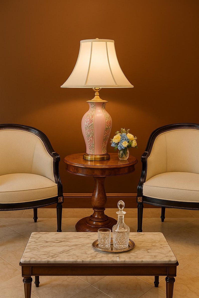 May include: A pink floral table lamp with a cream-colored lampshade sits on a small wooden table. Two cream-colored chairs with black trim flank the table. A marble-topped coffee table holds a decanter, two glasses, and a small tray.