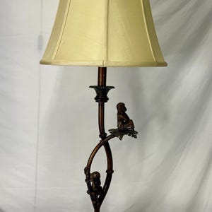 Puede incluir: Lámpara de mesa en tono bronce con pantalla beige. La lámpara presenta un diseño decorativo de monos trepando por una rama curvada. La base es redonda y detallada. La lámpara mide aproximadamente 71 cm de altura.