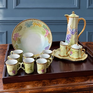 May include: A porcelain tea set featuring a large bowl, a tall pitcher, a creamer, a sugar bowl on a small tray, and six mugs. Each piece has a pale green base with painted purple grape clusters, green leaves, and gold-painted scalloped rims.