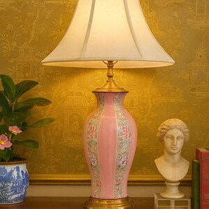May include: A pink ceramic table lamp with a cream-colored lampshade. The lamp has a floral design and sits on a gold base. A small statue and a potted plant are on the table.