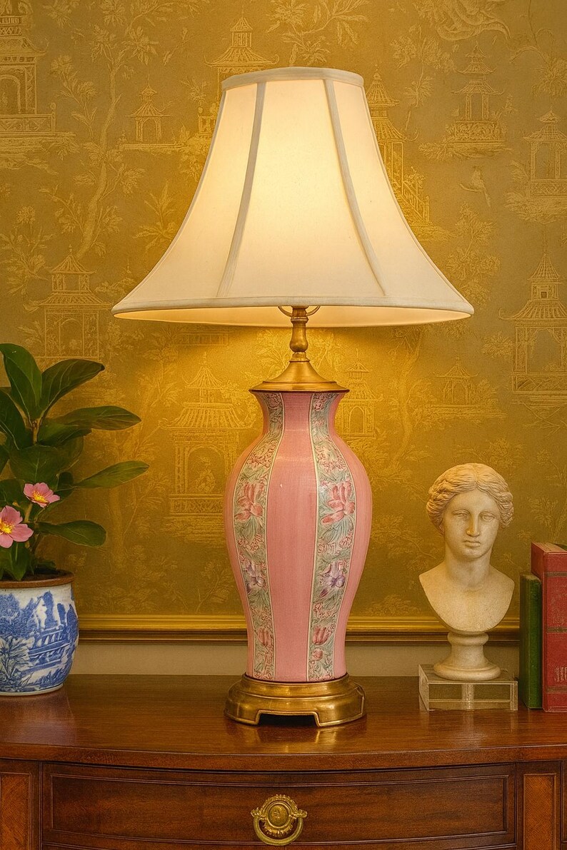 May include: A pink ceramic table lamp with a cream-colored lampshade. The lamp has a floral design and sits on a gold base. A small statue and a potted plant are on the table.