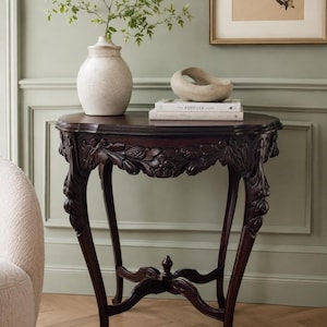 May include: A dark brown wooden console table with intricate floral carvings along the apron and cabriole legs. The table features a central stretcher with a finial and supports a speckled ceramic vase, a stone sculpture, and two stacked books.