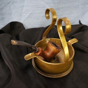 May include: A gold metal pipe stand with a wooden pipe resting inside. The stand has a round base and two curved arms that hold the pipe in place.