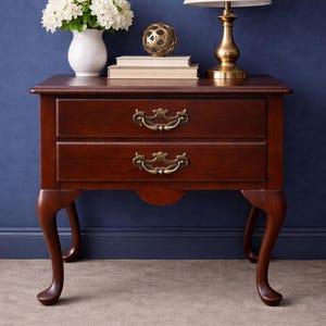May include: A dark cherry wood nightstand with two drawers and brass hardware. A white lamp with a cream shade sits on top, along with books, a decorative gold sphere, and a white vase with white flowers.