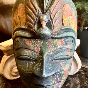 Può includere: Una maschera in legno intagliata a mano con dettagli intricati e colori vivaci. La maschera presenta un viso umano con gli occhi chiusi e un copricapo stilizzato. La maschera è dipinta in tonalità di blu, verde, giallo e rosso.