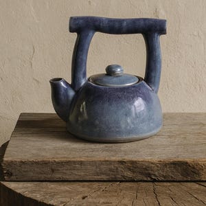 May include: A blue ceramic teapot with a rounded body, a curved handle, and a small spout. The teapot has a lid with a small knob and sits on a weathered wooden surface. The teapot's color transitions from a darker blue at the top to a lighter blue at the bottom.