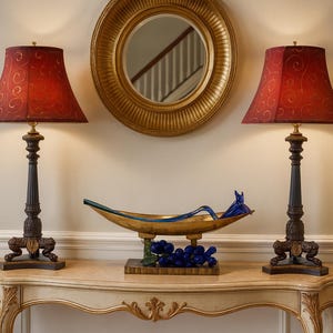 May include: Two ornate table lamps with red patterned shades flank a decorative gold-toned mirror. A gold and blue decorative bowl sits on a cream-colored table. The lamps have dark brown bases and are lit.