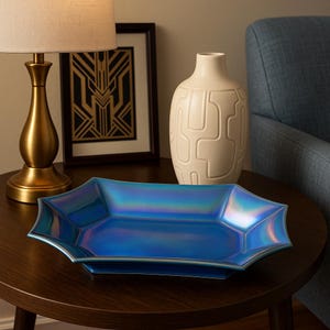 May include: A blue iridescent decorative tray with a starburst shape sits on a dark brown wooden table. A gold lamp and a white vase with a geometric pattern are also on the table. A framed art deco print is in the background.
