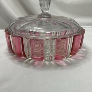 May include: A clear glass candy dish with a pink and clear ribbed design. The dish has a clear lid with a ribbed handle. The candy dish is likely vintage and is sitting on a white surface.