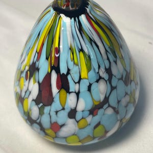 May include: A colorful, teardrop-shaped glass vase. The vase features a light blue base with a speckled pattern of white, yellow, red, and black. Vertical streaks of yellow, green, and white extend from the base to the black opening at the top.