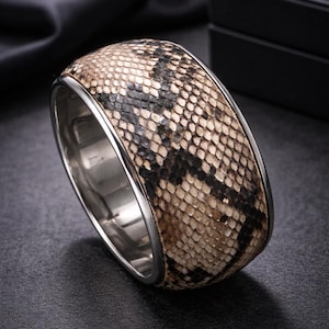May include: A wide, silver-toned bracelet with a snakeskin pattern. The bracelet features a band of genuine snakeskin in shades of brown, tan, and black, encased in a polished metal frame. The jewelry is displayed on a dark surface.