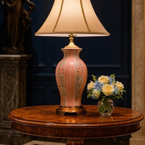 May include: A decorative table lamp with a pink ceramic base featuring floral designs, topped with a beige lampshade. The lamp sits on a round, wooden table, next to a small vase of flowers. The room has a dark blue wall.