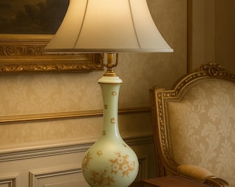 May include: A vintage-style table lamp with a cream-colored lampshade and a pale green base adorned with gold floral patterns. The lamp sits on a dark wooden table next to two stacked books. The lamp's design evokes a classic, elegant aesthetic.