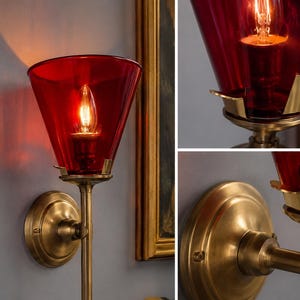 May include: A wall sconce with a red glass shade and a brass finish. The sconce has a conical shade and a decorative brass base. The light bulb is visible inside the shade, illuminating the area. The sconce is mounted on a grey wall.
