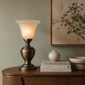 May include: A table lamp with a dark bronze-toned metal base and a frosted glass shade shaped like an open flower. The lamp is illuminated, casting a soft glow. Beside it are three stacked books titled LIVE BEAUTIFULLY, at 100, and THIS IS HOME.