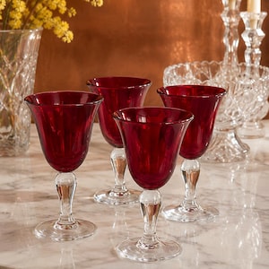 May include: A set of four wine glasses featuring deep red bowls and clear glass stems with circular bases. The glasses have a smooth, curved silhouette and are arranged in a group, showcasing the contrast between the colored glass and clear stems.