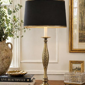 May include: A decorative table lamp with a black lampshade and a gold-colored base. The lamp has an ornate, detailed design and sits on a wooden surface. The lamp is next to a window and other decorative items.