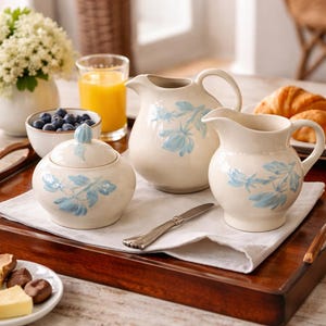 May include: A cream-coloured ceramic breakfast set featuring a sugar bowl with a lid, a small jug, and a larger jug, all adorned with blue floral designs. The set is arranged on a wooden tray with a white napkin, alongside a knife.