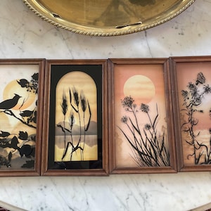 May include: Four framed silhouette art pieces with wooden frames. Each frame features a different nature scene: a bird on a branch, wheat stalks, a sunset with tall grass, and a sunset with a butterfly on a plant. The backgrounds have warm orange and yellow tones.