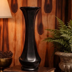 May include: A tall, black vase with a flared top and a rounded base. The vase is made of a glossy material and sits on top of a stack of old books. The background features a wooden wall and a potted fern.