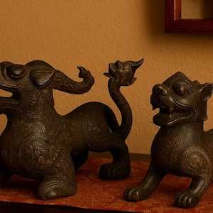 May include: Two bronze-colored mythical creature statues. The statues have detailed features, including open mouths, and decorative patterns. One statue has a long, curved neck and head. The other statue is in a seated position.