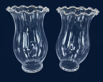May include: Two clear glass lamp shades with a ruffled edge. The shades have a flared shape and a smooth, transparent surface. The shades are designed to fit over a light bulb and diffuse light.