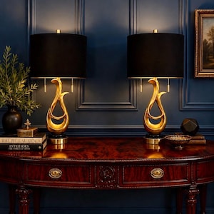 May include: A pair of table lamps featuring gold-toned, abstract, fluid-shaped bases with black cylindrical shades. Each lamp has two pull chains for operation and sits on a circular black base, providing a symmetrical lighting arrangement.