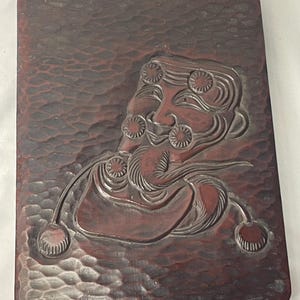 May include: A rectangular, reddish-brown wooden tray with a carved relief of a stylized face with floral accents. The tray has a textured surface and rounded corners, showcasing a traditional design.