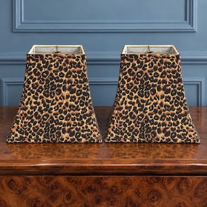 May include: A pair of matching lamp shades featuring a classic leopard print pattern in shades of black, brown, and tan. Each shade has a square, tapered silhouette with a metal spider fitter at the top, showcasing a structured fabric design.