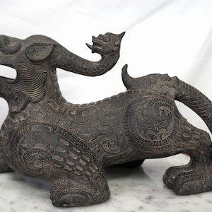 May include: A dark bronze statue of a mythical creature, featuring an open mouth, horns, and detailed patterns. The sculpture is displayed on a white marble surface, highlighting its intricate design and craftsmanship.