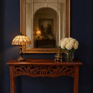 May include: A decorative entryway scene featuring a gold-framed mirror above a wooden console table. The table holds a stained glass lamp, a small decorative box, and a vase of white flowers. The wall is painted a deep blue.