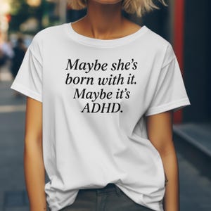 May include: White t-shirt featuring the text "Maybe she's born with it. Maybe it's ADHD" in black, elegant lettering. The shirt has a round neck and short sleeves. The image is taken outside.