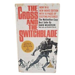 May include: A vintage paperback book cover with a red and white title "The Cross and the Switchblade". The cover features a black and white illustration of a man running with a knife in his hand. The text "Now in a new movie edition with 16 pages of exciting pictures!" is printed in red and white.