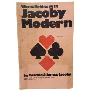 May include: A softcover book titled Win at Bridge with Jacoby Modern by Oswald & James Jacoby. The cover is light pink with large black text and a central graphic featuring a red heart, a black spade, a black club, and a red diamond symbol.