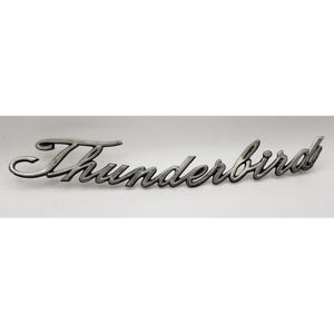 May include: A silver-colored Thunderbird emblem, featuring the word "Thunderbird" in a cursive script. The emblem has a slightly weathered appearance, with a hint of dark shading. The background is plain white.
