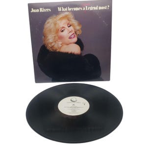 May include: A vinyl record and album cover featuring Joan Rivers. The album cover has a photo of Rivers with blonde hair and a black fur wrap. The album title is "What becomes a Legend most?" The record is black with a white label.