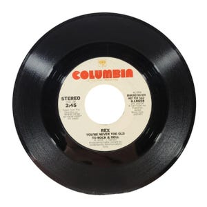 May include: A black vinyl record with a white label. The label has the text "Columbia" in red, "Stereo" in black, "Intro. Of 2:45" in black, "Rex You're Never Too Old To Rock & Roll" in black, and "45 RPM Demonstration Not For Sale 3-10658" in black.