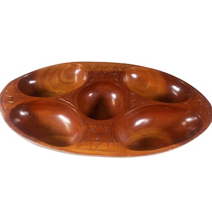 May include: A wooden serving tray with six oval-shaped compartments. The tray is made of polished, dark brown wood with carved decorative details between the compartments. The compartments are designed to hold various snacks or appetizers.