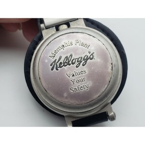 May include: A silver-toned watch face with the Kellogg's logo and the text "Memphis Plant Values Your Safety." The watch has a black leather band and a silver metal clasp.