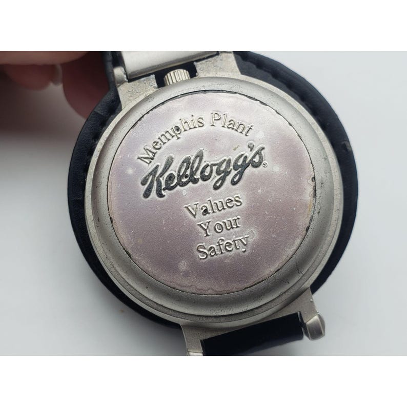 May include: A silver-toned watch face with the Kellogg's logo and the text "Memphis Plant Values Your Safety." The watch has a black leather band and a silver metal clasp.