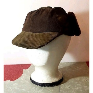 May include: A brown corduroy cap with a rounded crown and a contrasting olive green brim. The cap has ear flaps and is displayed on a white mannequin head. The hat is made of a ribbed fabric.