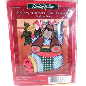 May include: Holiday "Greeters" plastic canvas kit featuring a stocking bear design. The kit includes a brown bear with a blue scarf, sitting in a Christmas stocking. The finished size is 10 x 13 inches.
