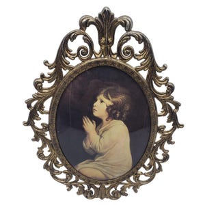 May include: An oval-shaped, antique gold-colored picture frame with an ornate, scrolling design. The frame holds a print of a child with curly hair, hands clasped in prayer, set against a dark background. A vintage religious-themed item.