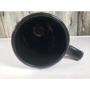 May include: A black ceramic mug with a handle, viewed from above. The interior of the mug is dark, and the handle is on the right side. The mug is sitting on a white surface.