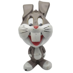 May include: A vintage Bugs Bunny figurine. The figure is primarily gray and white, with a red nose and mouth. The cartoon character is wearing a gray suit and has large, expressive eyes. The figurine has visible signs of wear.