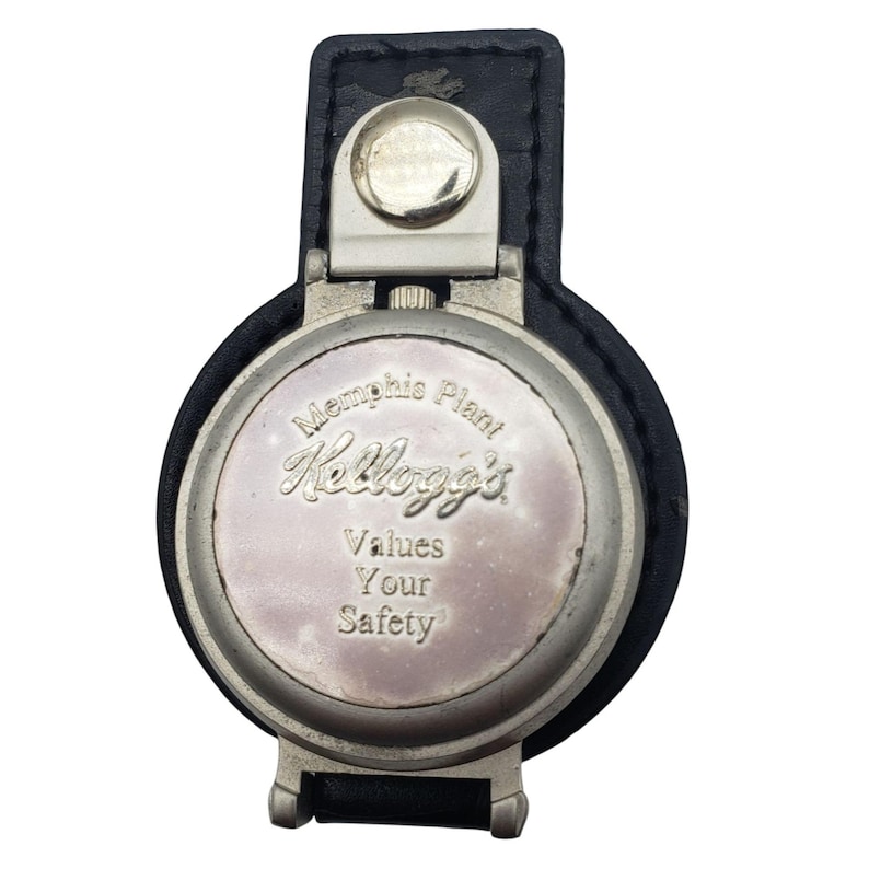 May include: A vintage silver-toned metal fob with a black leather strap. The fob features the Kellogg's logo and the text "Memphis Plant Values Your Safety". The fob has a circular design and a hinged clasp.