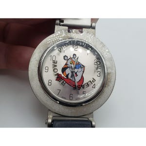 May include: A silver-toned wristwatch with a round face featuring a cartoon tiger character. The watch face has the words "REMOVE FOIL PLEASE" and the numbers 1 through 12. The watch has a silver band and a dark blue leather strap.